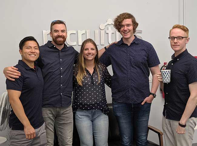 Recruitics employees embrace for a photo in front of the Recruitics logo